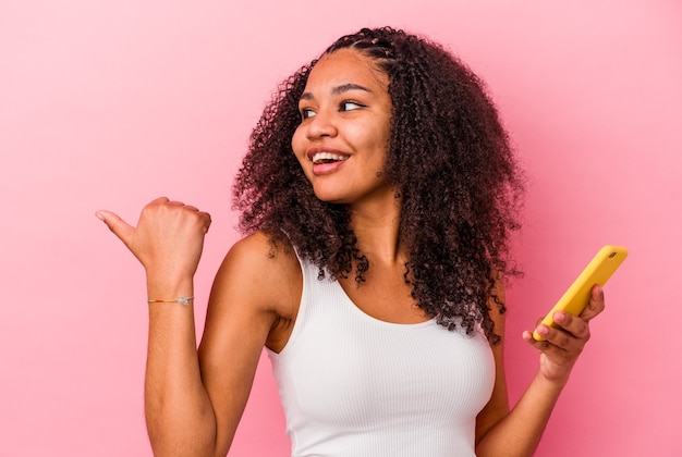 Junge Afroamerikanerfrau, die ein Mobiltelefon hält, zeigt mit Daumenfinger weg, lachend