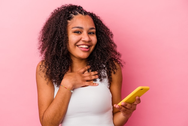 Junge Afroamerikanerfrau, die ein Mobiltelefon lokalisiert einen rosa Hintergrund hält, lacht laut heraus und hält Hand auf Brust.