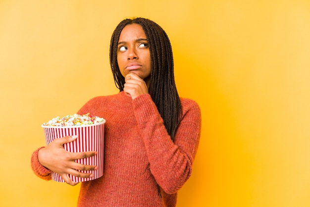 Junge Afroamerikanerfrau, die ein Popcorn lokalisiert hält, das seitwärts mit zweifelhaftem und skeptischem Ausdruck schaut.