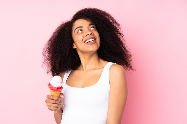 Junge afroamerikanerfrau, die eine kornett-eiscreme lokalisiert auf rosa hält, die beim lächeln oben betrachtet