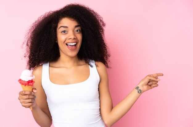 Junge Afroamerikanerfrau, die eine Kornett-Eiscreme lokalisiert auf rosa überrascht hält und Finger zur Seite zeigt