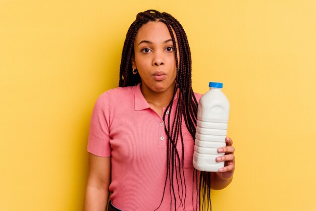 Junge Afroamerikanerfrau, die eine Milchflasche lokalisiert auf gelber Wand zuckt, zuckt mit den Schultern und offenen Augen verwirrt.