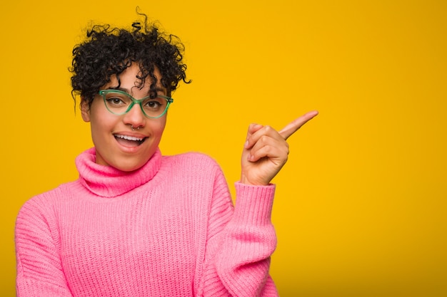 Junge Afroamerikanerfrau, die eine rosa Strickjacke lächelt trägt, freundlich zeigend mit dem Zeigefinger weg.