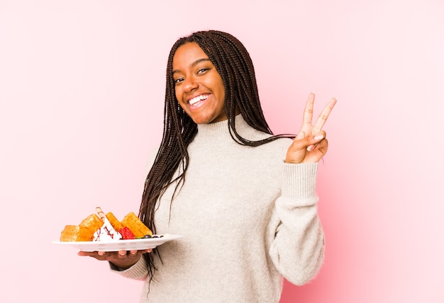 Junge Afroamerikanerfrau, die eine Waffel isoliert freudig und sorglos hält, ein Friedenssymbol mit den Fingern zeigend.