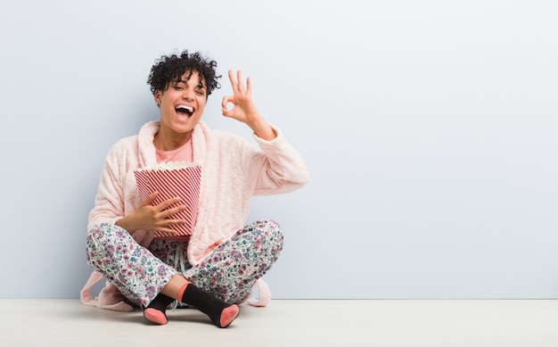 Junge Afroamerikanerfrau, die einen Popcorn-Eimer hält, der fröhlich und zuversichtlich zeigt ok ok Geste.