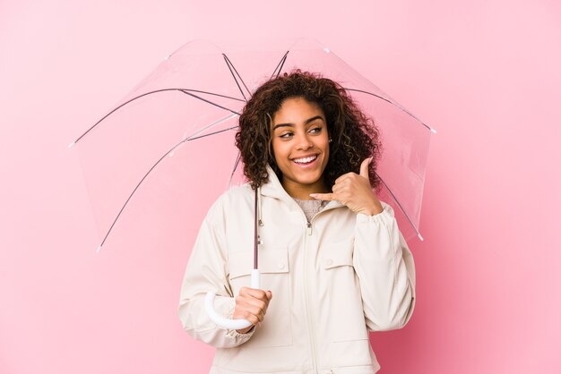 Junge Afroamerikanerfrau, die einen Regenschirm hält, der eine Handy-Anrufgeste mit den Fingern zeigt.