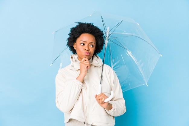 Junge Afroamerikanerfrau, die einen Regenschirm hält, der seitwärts mit zweifelhaftem und skeptischem Ausdruck schaut.