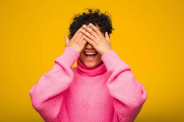 Junge Afroamerikanerfrau, die einen rosa Pullover mit den Augen der Hände trägt, lächelt breit und wartet auf eine Überraschung.