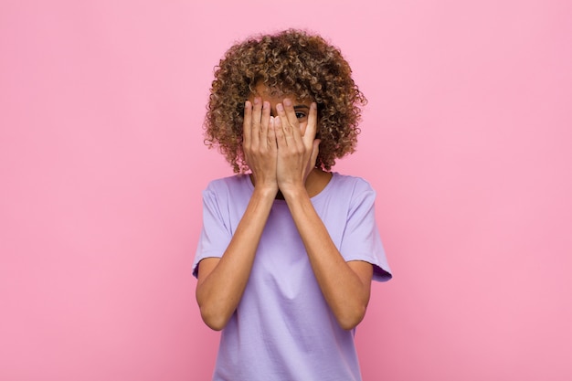 Junge Afroamerikanerfrau, die Gesicht mit Händen bedeckt, zwischen Fingern mit überraschtem Ausdruck späht und zur Seite gegen rosa Wand schaut