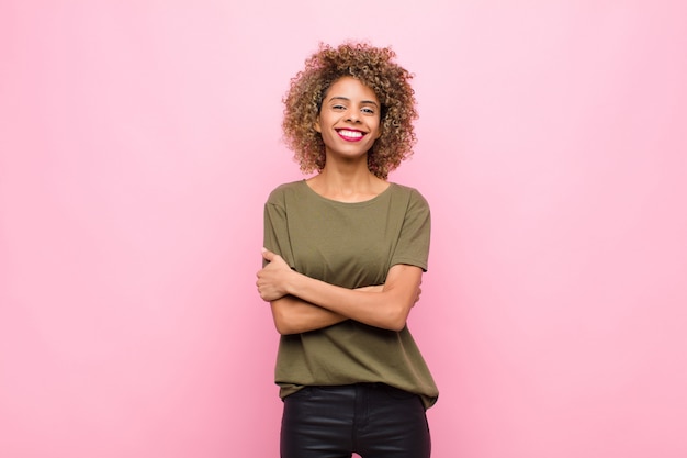 Junge Afroamerikanerfrau, die glücklich mit verschränkten Armen lacht, mit einer entspannten, positiven und zufriedenen Haltung gegen rosa Wand
