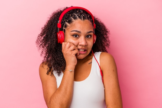 Junge Afroamerikanerfrau, die Musik mit Kopfhörern lokalisiert auf rosa Hintergrund beißenden Fingernägeln, nervös und sehr ängstlich hört.