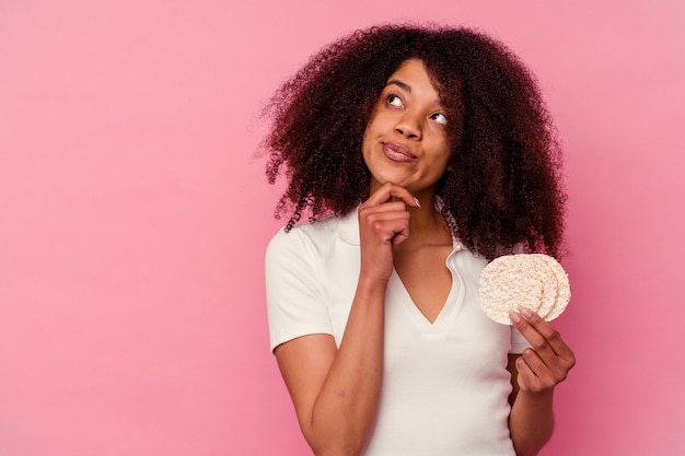 Junge Afroamerikanerfrau, die Reiskuchen isst lokalisiert auf rosa Hintergrund, der seitwärts mit zweifelhaftem und skeptischem Ausdruck schaut.