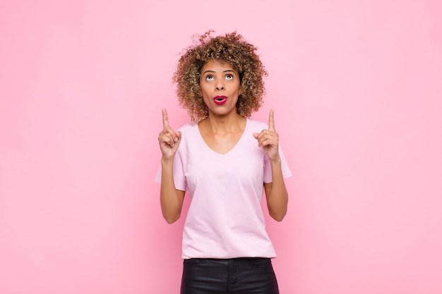 Junge Afroamerikanerfrau, die sich ehrfürchtig und mit offenem Mund nach oben zeigend mit einem schockierten und überraschten Blick auf rosa Wand fühlt