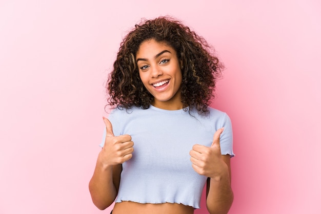 Junge Afroamerikanerfrau gegen eine rosa Oberfläche, die beide Daumen anhebt, lächelnd und zuversichtlich.