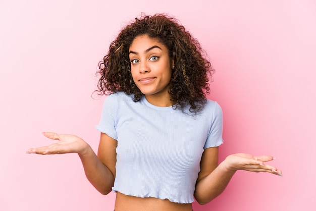 Junge Afroamerikanerfrau gegen eine rosa Wand, die Schultern in fragender Geste zweifelt und zuckt.