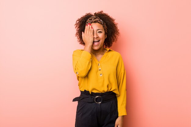 Junge Afroamerikanerfrau gegen eine rosa Wand, die Spaß hat, die Hälfte des Gesichtes mit Palme zu bedecken.