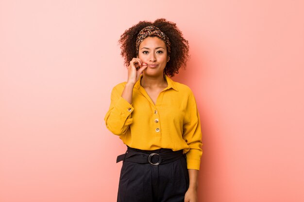 Junge afroamerikanerfrau gegen eine rosa wand mit den fingern auf den lippen, die ein geheimnis halten.