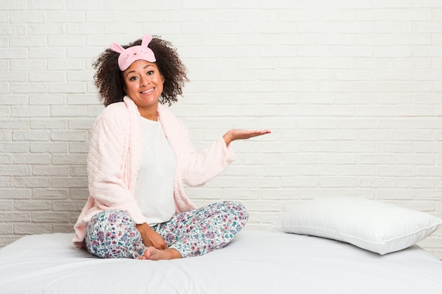 Junge Afroamerikanerfrau im tragenden pijama des Betts, das einen Kopienraum auf einer Palme zeigt und eine andere Hand auf Taille hält.