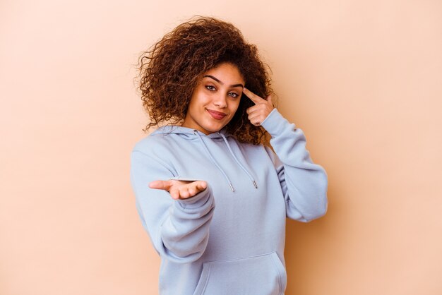 Junge Afroamerikanerfrau lokalisiert auf beigem Hintergrund, der ein Produkt an der Hand hält und zeigt.