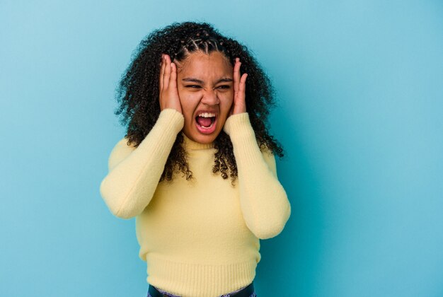 Junge Afroamerikanerfrau lokalisiert auf blauen Wand, die Ohren mit Händen bedeckt, die versuchen, nicht zu lautes Geräusch zu hören.