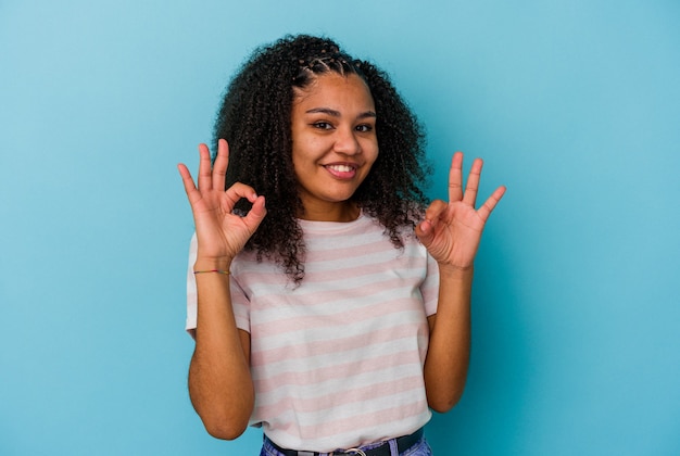 Junge Afroamerikanerfrau lokalisiert auf blauer Wand fröhlich und zuversichtlich, ok Geste zeigend.