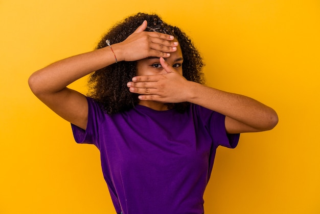 Junge Afroamerikanerfrau lokalisiert auf gelbem Hintergrund blinken an der Kamera durch Finger, verlegenes bedeckendes Gesicht.