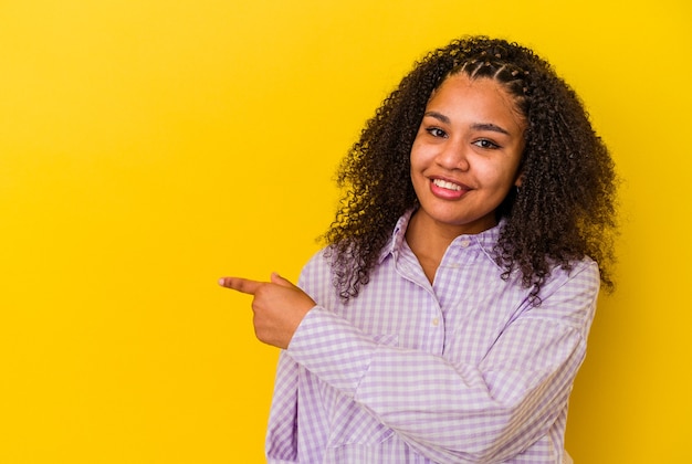 Junge Afroamerikanerfrau lokalisiert auf gelber Wand, die lächelt und beiseite zeigt und etwas an der leeren Stelle zeigt.