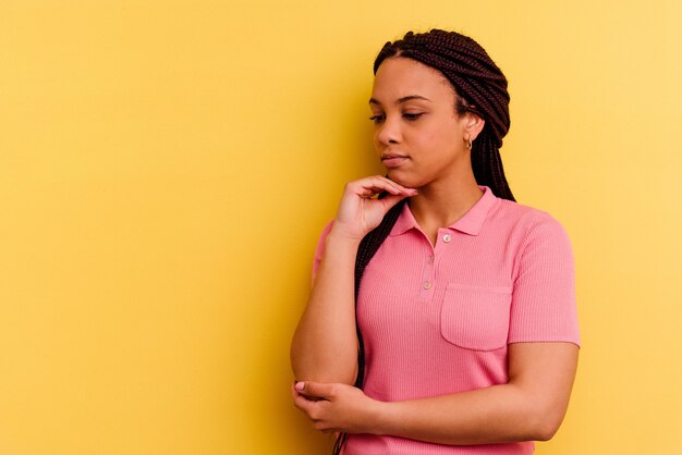 Junge Afroamerikanerfrau lokalisiert auf gelber Wand, die seitwärts mit zweifelhaftem und skeptischem Ausdruck schaut.