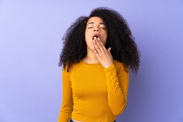 Junge Afroamerikanerfrau lokalisiert auf lila Gähnen und Kegeln weit offenem Mund mit Hand