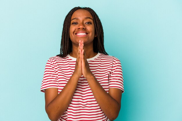 Junge Afroamerikanerin, die auf blauem Hintergrund isoliert ist und die Hände im Gebet in der Nähe des Mundes hält, fühlt sich zuversichtlich.