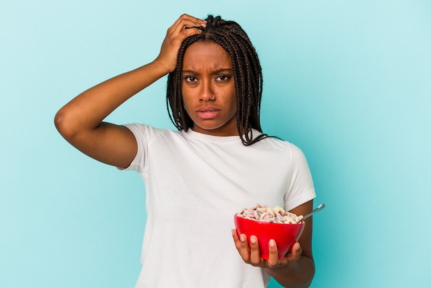 Junge Afroamerikanerin, die eine Schüssel mit Cerealien isoliert auf blauem Hintergrund hält und schockiert ist, hat sich an ein wichtiges Treffen erinnert.