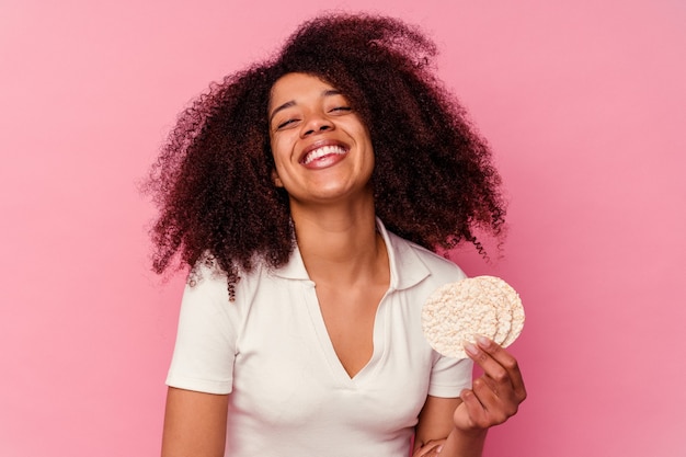 Junge Afroamerikanerin, die einen Reiskuchen isst, der auf rosafarbenem Hintergrund lokalisiert wird, lacht und Spaß hat.