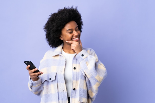 Junge Afroamerikanerin, die Handy verwendet, lokalisiert auf lila, das eine Idee denkt und Seite schaut