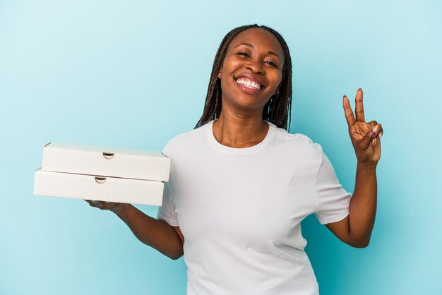 Junge afroamerikanerin, die pizzas einzeln auf blauem hintergrund hält, fröhlich und sorglos, die ein friedenssymbol mit den fingern zeigen.