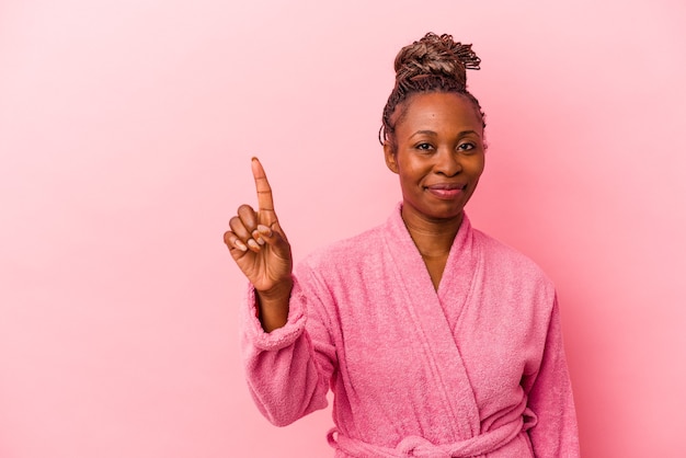 Junge Afroamerikanerin, die rosafarbenen Bademantel trägt, einzeln auf rosafarbenem Hintergrund, der Nummer eins mit dem Finger zeigt.