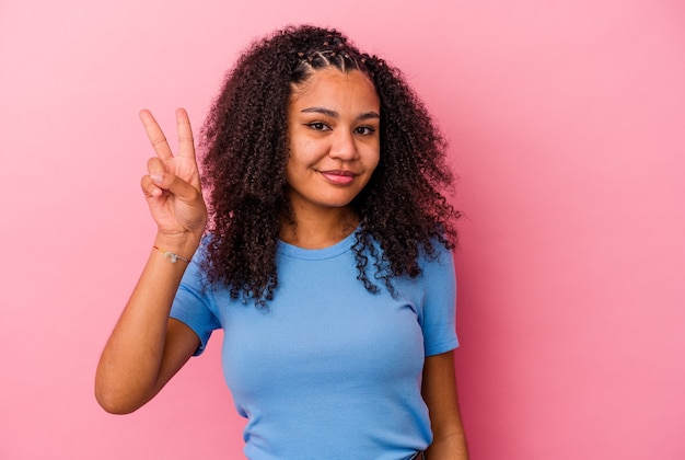 Junge Afroamerikanerin einzeln auf rosafarbenem Hintergrund fröhlich und sorglos, die ein Friedenssymbol mit den Fingern zeigt.