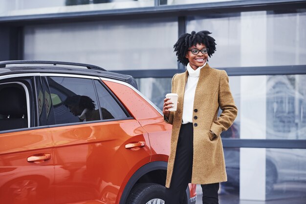 Junge Afroamerikanerin in Gläsern und mit einer Tasse Getränk steht draußen in der Nähe eines modernen Autos.