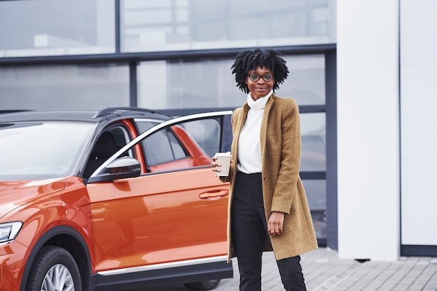 Junge Afroamerikanerin in Gläsern und mit einer Tasse Getränk steht draußen in der Nähe eines modernen Autos.