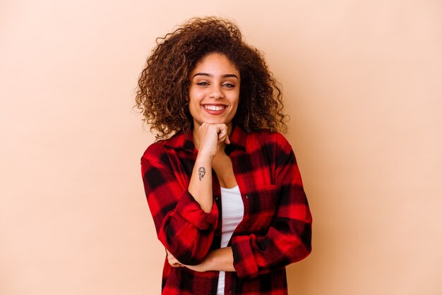 Junge Afroamerikanerin isoliert auf beigefarbenem Hintergrund, die glücklich und selbstbewusst lächelt und das Kinn mit der Hand berührt.