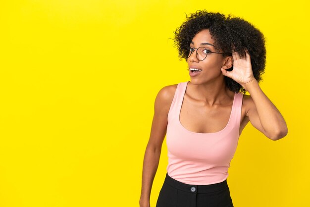 Junge Afroamerikanerin isoliert auf gelbem Hintergrund, die etwas hört, indem sie die Hand auf das Ohr legt