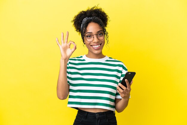 Junge Afroamerikanerin isoliert auf gelbem Hintergrund mit Handy und macht OK-Zeichen