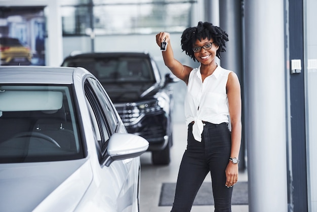 Junge Afroamerikanerin mit Brille steht im Autosalon in der Nähe des Fahrzeugs mit Schlüsseln in den Händen.