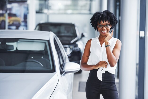 Junge Afroamerikanerin mit Brille steht im Autosalon in der Nähe des Fahrzeugs.