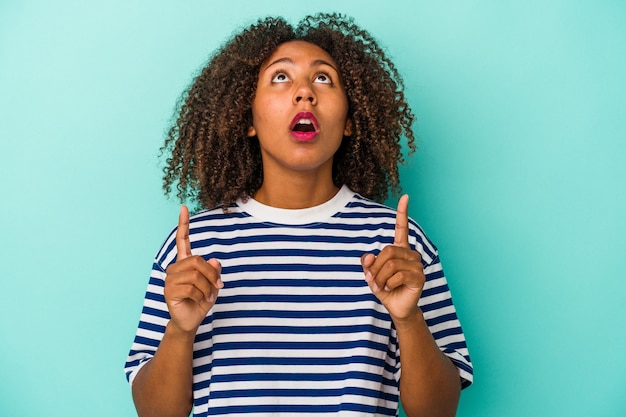 Junge Afroamerikanerin mit lockigem Haar auf blauem Hintergrund isoliert, die mit geöffnetem Mund nach oben zeigt.