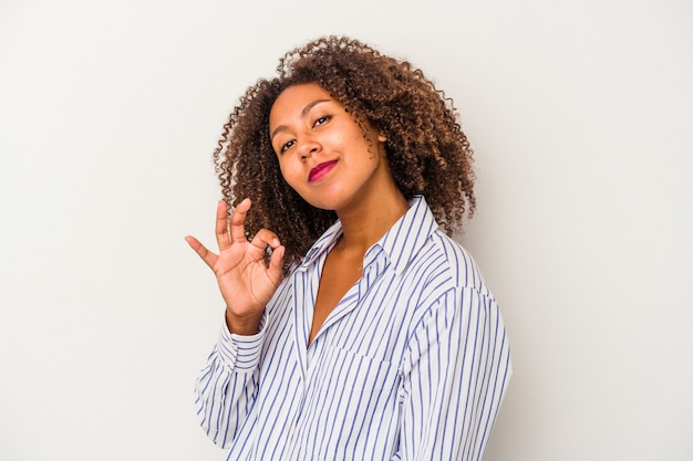 Junge Afroamerikanerin mit lockigem Haar, die auf weißem Hintergrund isoliert ist, zwinkert ein Auge und hält eine okaygeste mit der Hand.