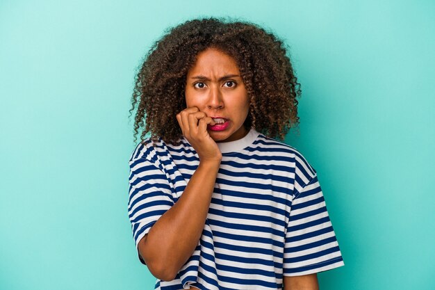 Junge Afroamerikanerin mit lockigem Haar einzeln auf blauem Hintergrund beißende Fingernägel, nervös und sehr ängstlich.