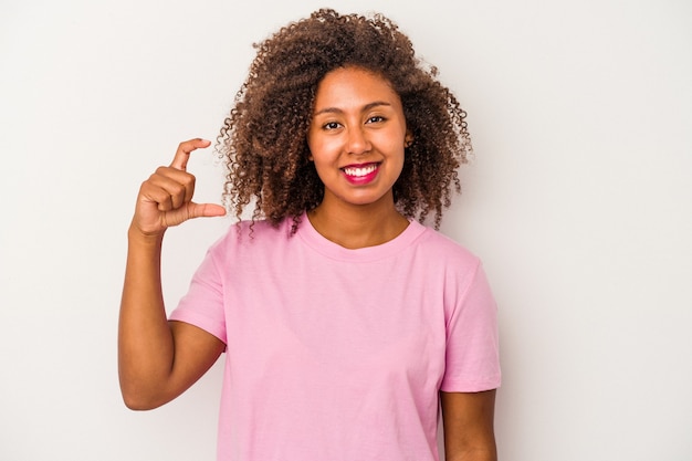 Junge Afroamerikanerin mit lockigem Haar isoliert auf weißem Hintergrund, die etwas Kleines mit den Zeigefingern hält, lächelnd und selbstbewusst.