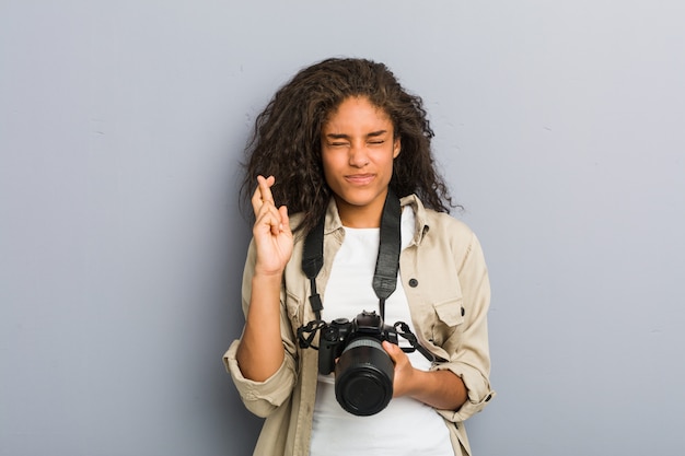 Junge Afroamerikanerphotographfrau, die Finger einer Kameraüberfahrt für das Haben des Glücks hält