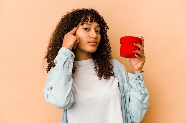 Junge afroamerikanische Afro-Frau, die ein Valentinstaggeschenk hält, das Tempel mit Finger zeigt, denkend, auf eine Aufgabe fokussiert.