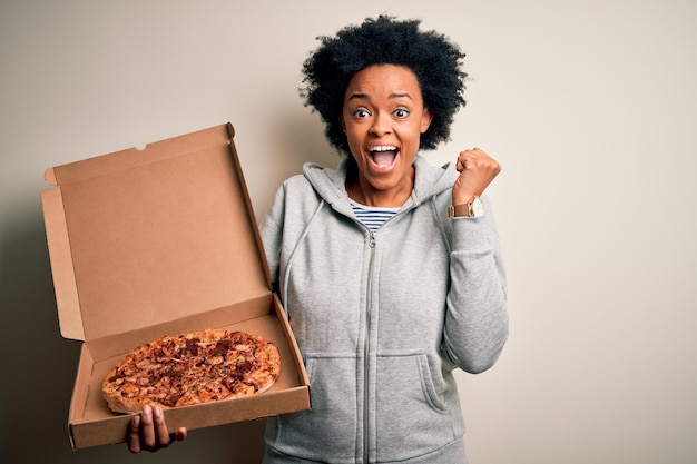 Junge afroamerikanische Afro-Frau mit lockigem Haar hält einen Lieferkarton mit italienischer Pizza in der Hand, schreit stolz und feiert Sieg und Erfolg, sehr aufgeregt und jubelnd